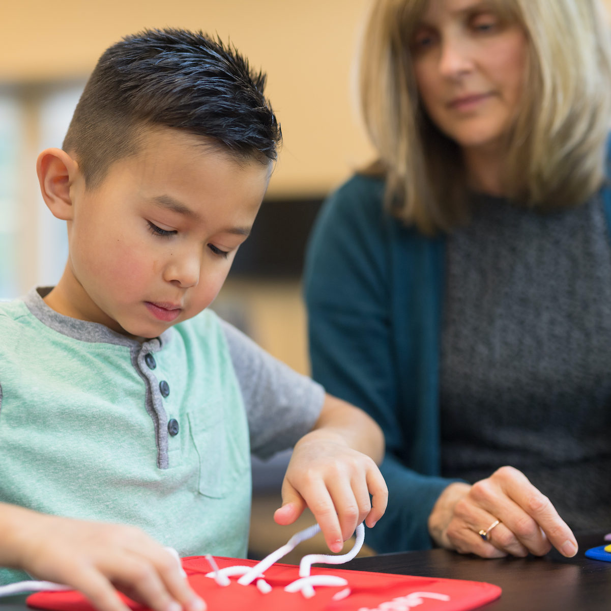 An occupational therapist works with a young elementary-age ethnic boy on his coordination skills. She is showing him a fun exercise for him to practice tying knots. The shot is focused on him.